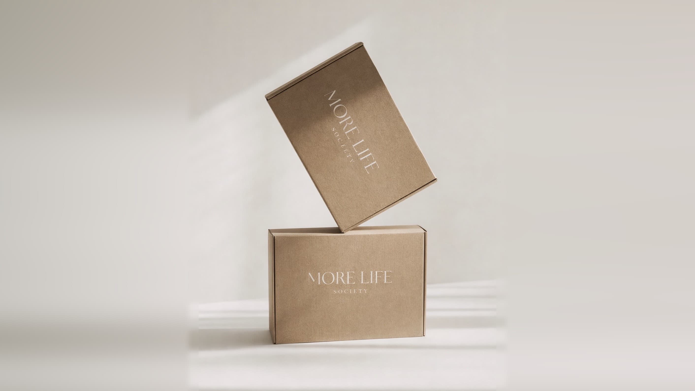 Two MoreLife Society mailer boxes in kraft board, one tilted against the other, the wordmark printed on each