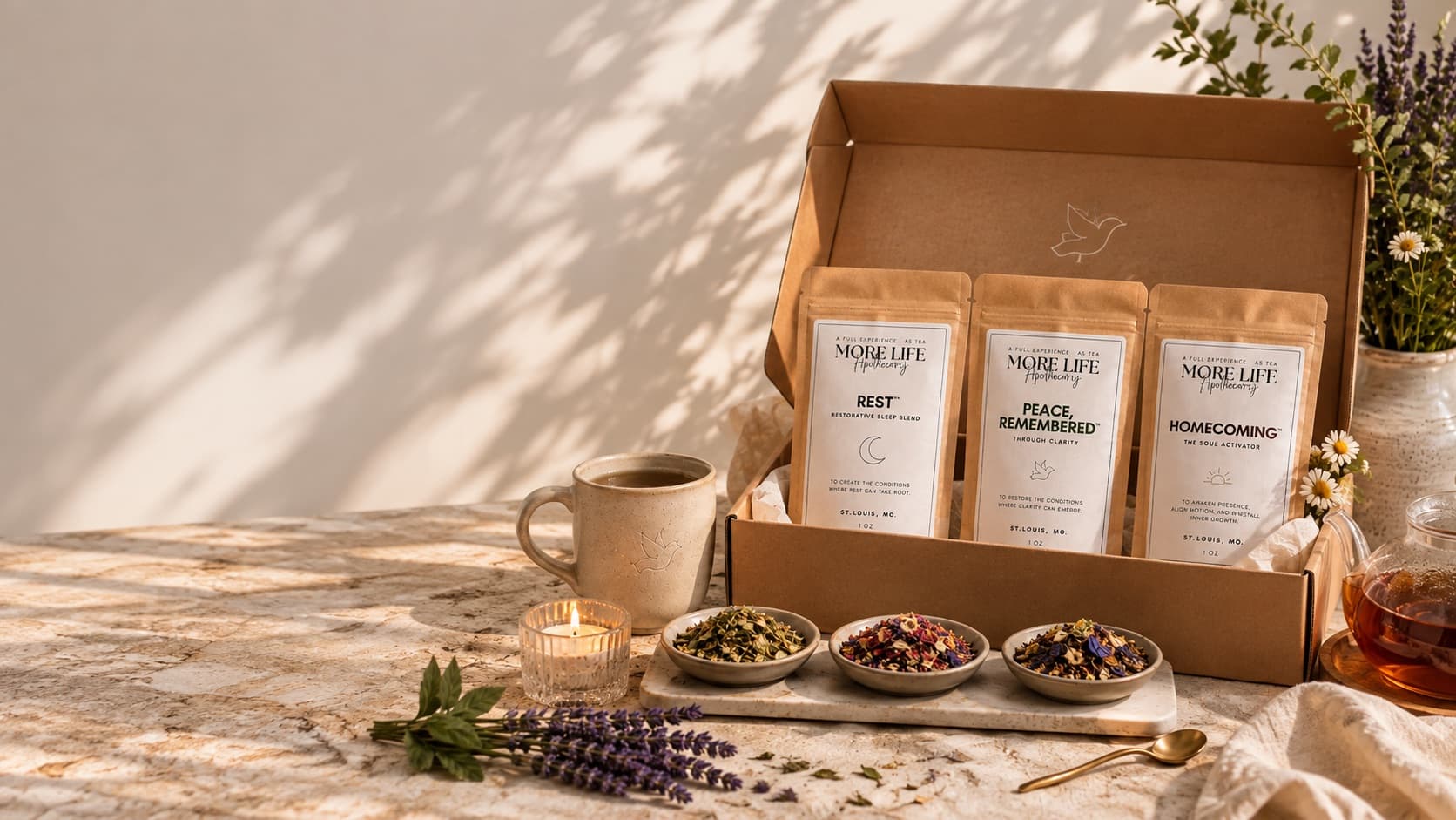 An open MoreLife Experience Box holding three pouches — Rest, Peace, Remembered and Homecoming — beside a mug, a candle and three bowls of loose tea in afternoon light