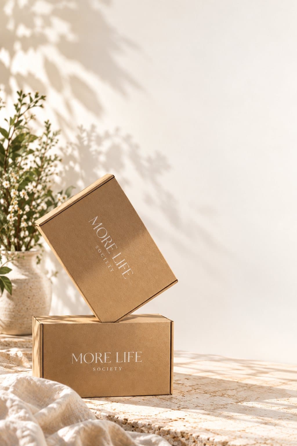 Two MoreLife Society boxes, one tilted on the other, beside a vase of white flowers in warm morning light