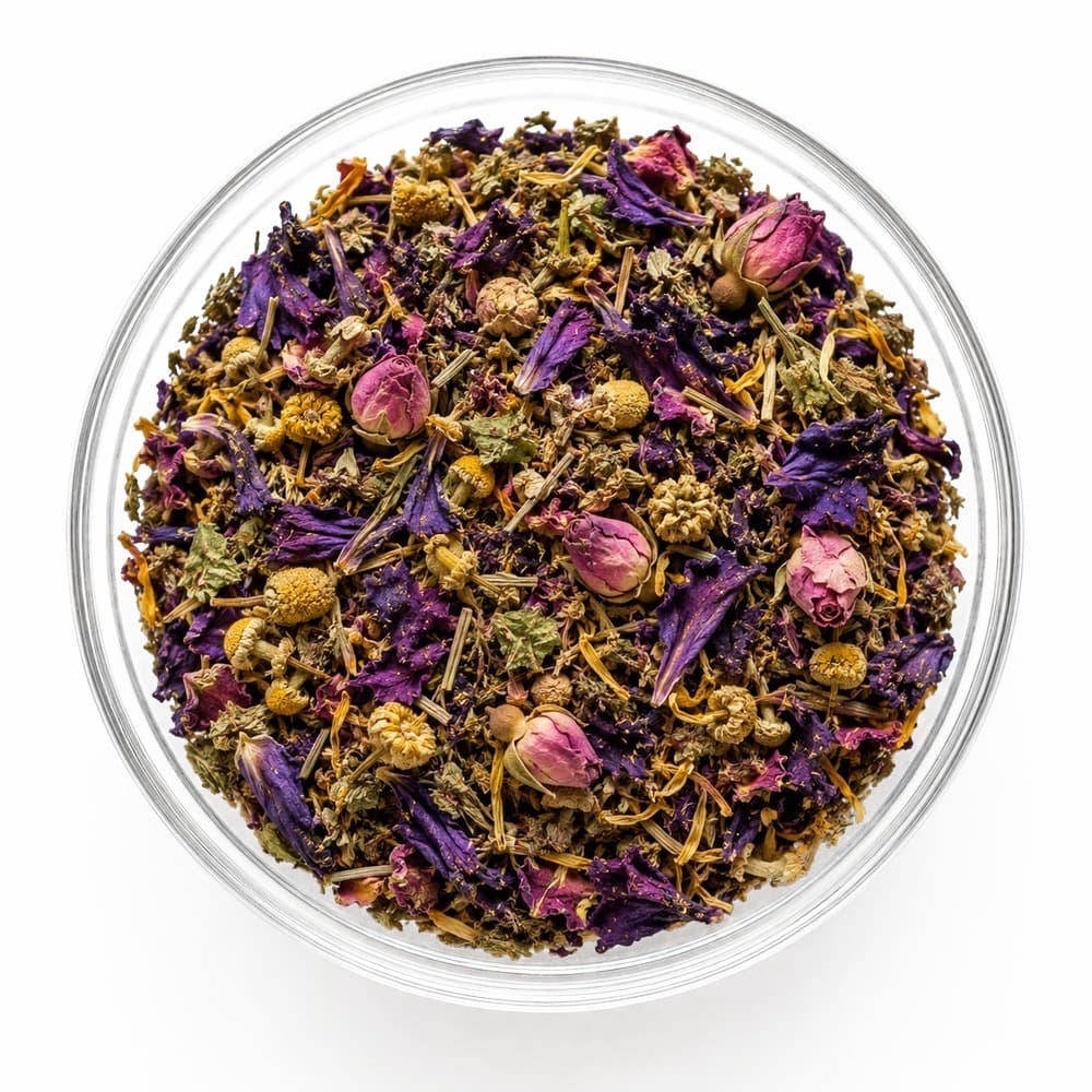 A glass bowl of loose-leaf Peace, Remembered tea — dried chamomile, calendula, violet and butterfly pea petals, and pink rosebuds, on a plain white background
