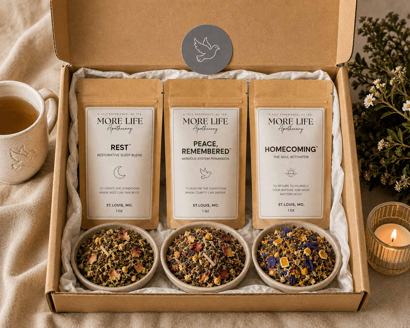 An open MoreLife Experience Box with three tea pouches — Rest, Peace Remembered, and Homecoming — each beside a bowl of its loose-leaf botanicals, with a cup of tea, candle, and flowers nearby