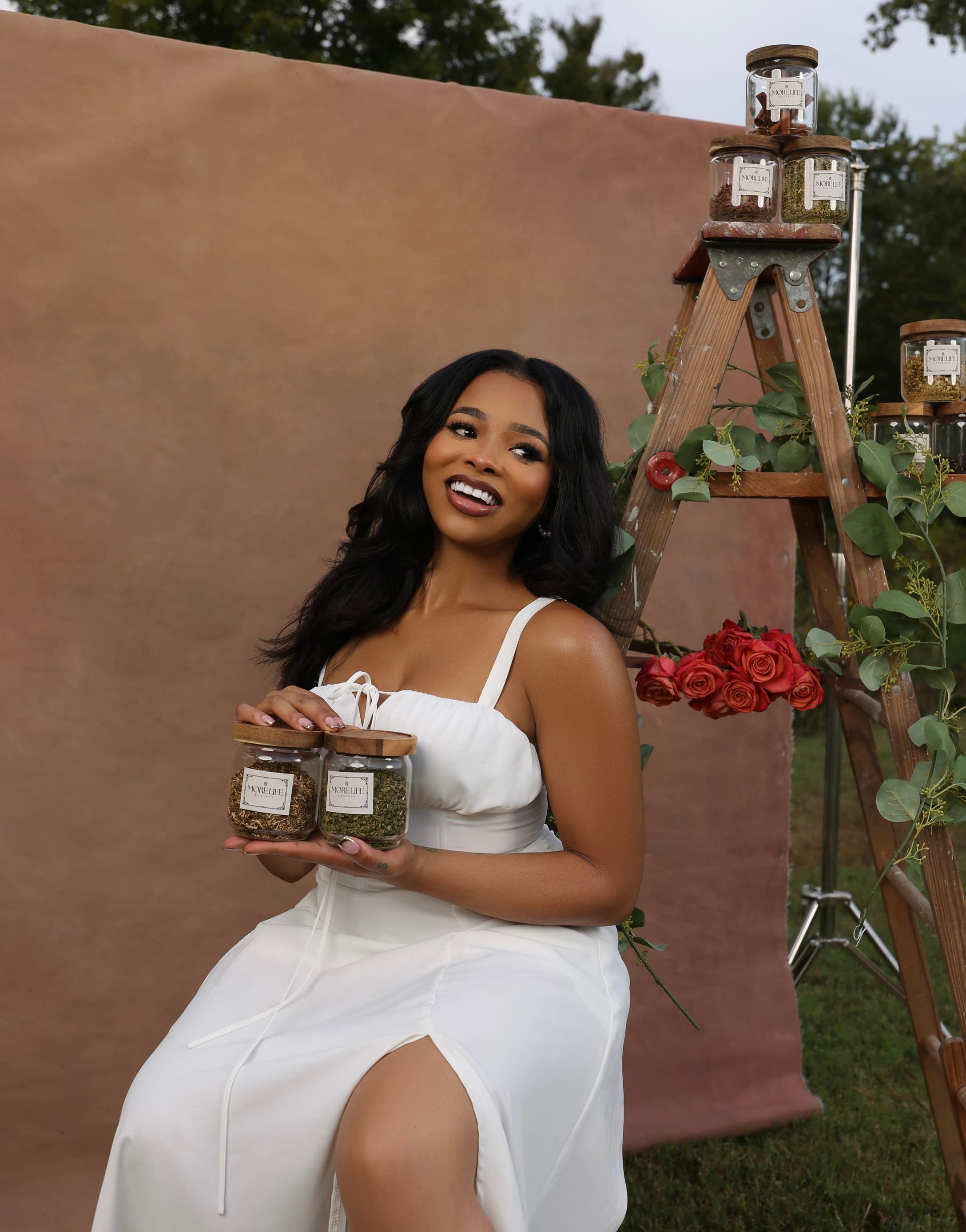 A woman smiling and holding two jars of MoreLife loose-leaf tea blends outdoors, with more jars, eucalyptus, and roses arranged on a wooden ladder behind her.